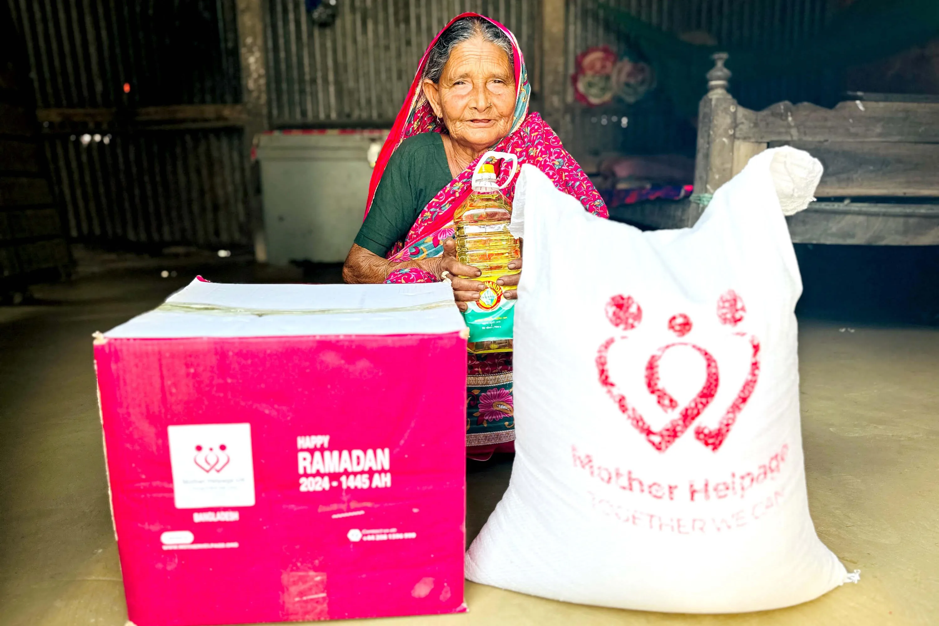 Elderly woman holding cooking oil beside a Mother Helpage Ramadan aid box and rice sack