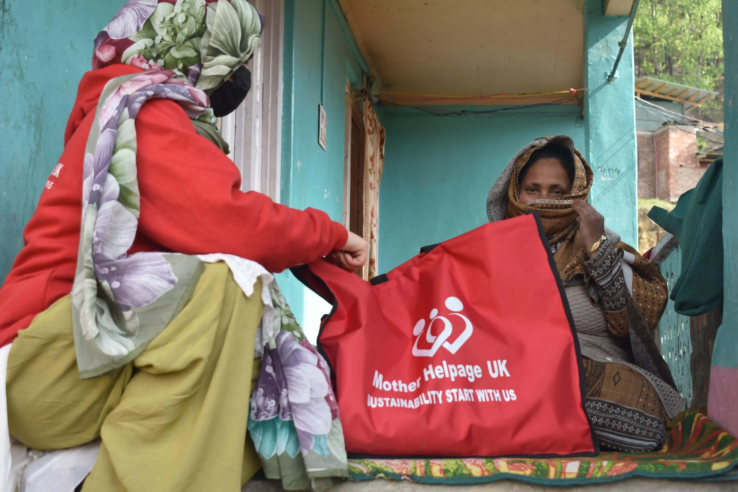 Mother Helpage volunteer handing a red Ramadan aid bag to an elderly woman on a covered porch
